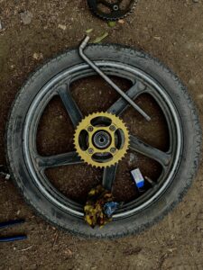Motorcycle wheel with sprocket and tools on dirt ground in DIY repair setup.