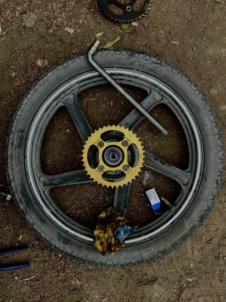 Motorcycle wheel with sprocket and tools on dirt ground in DIY repair setup.