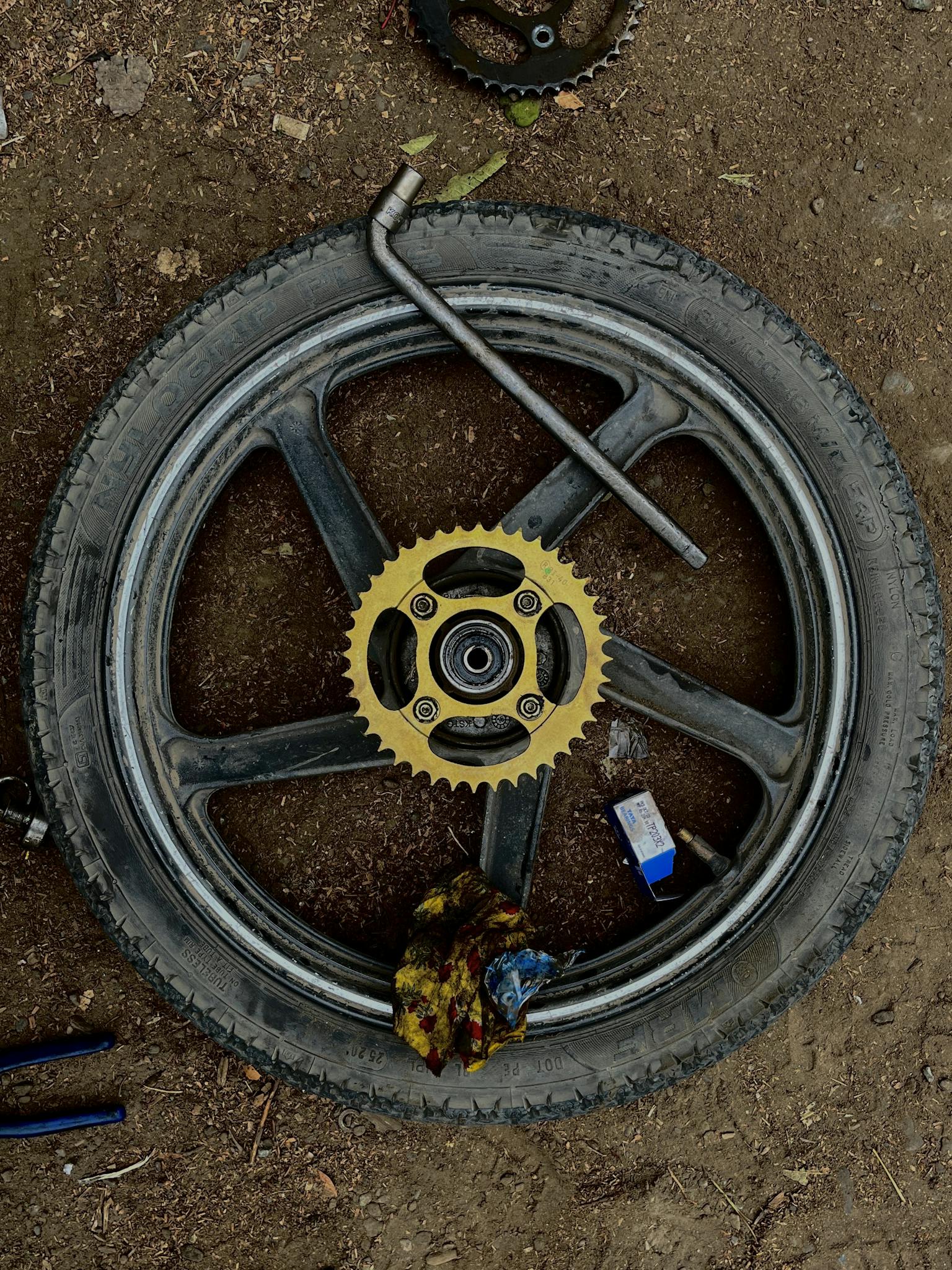 Motorcycle wheel with sprocket and tools on dirt ground in DIY repair setup.
